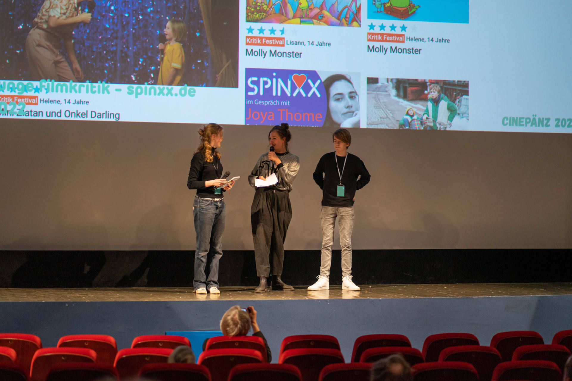 Festivalredaktion Mitglieder Lisann und Caspar stehen auf der Bühne des Odeon Kino Köln mit Christine Bernau, Festivalleitung des Filmfestival für junges Publikum - CINEPÄNZ
